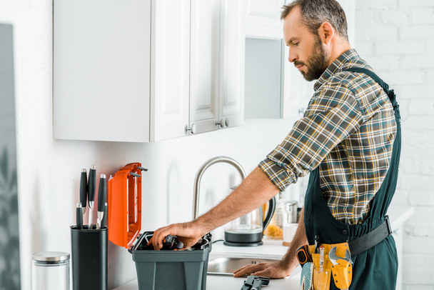 side view of handsome plumber taking tools from toolbox in kitchen - Fotoğraf, Görsel