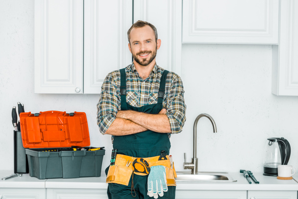 улыбающийся красивый сантехник, стоящий со скрещенными руками и смотрящий в камеру на кухне
 - Фото, изображение