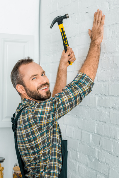 Улыбающийся красивый ремонтник забивает гвоздь в белую стену и смотрит в камеру
 - Фото, изображение