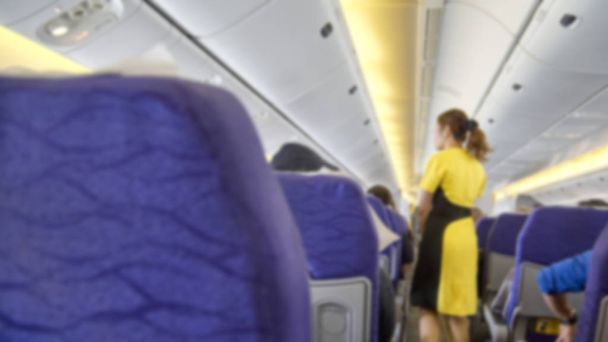 blurred Interior of commercial airplane with flight attandant serving passengers on seats during flight. Stewardess in dark Yellow uniform walking the aisle. Horizontal composition. - Photo, Image