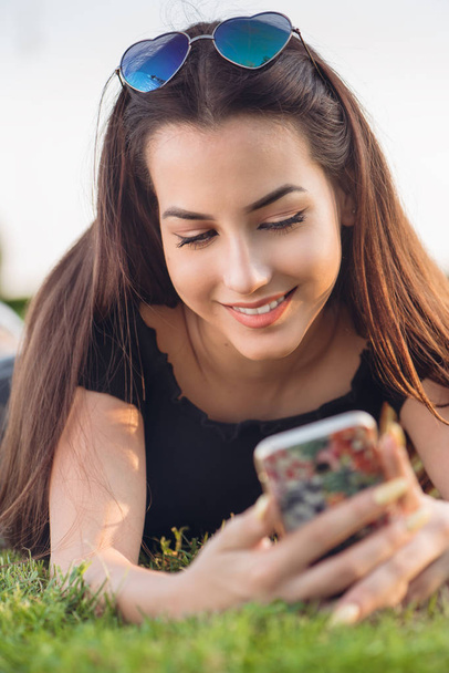 Chica sonriendo usando un teléfono inteligente acostado en la hierba
 - Foto, imagen