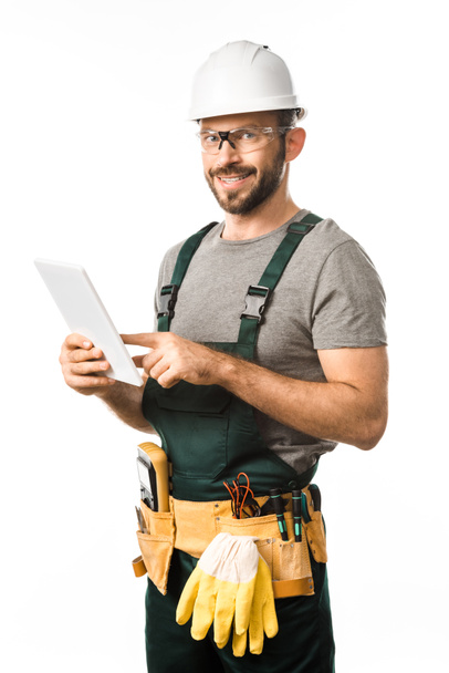 electricista guapo sonriente en casco y gafas protectoras usando tableta aislada en blanco
 - Foto, Imagen