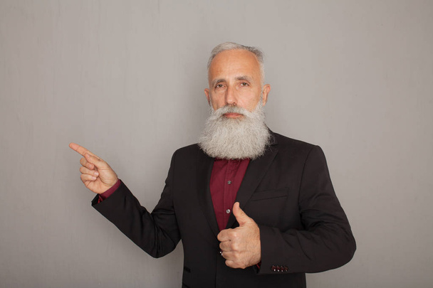Portrait of a happy old bearded businessman pointing finger away over grey background - Photo, image