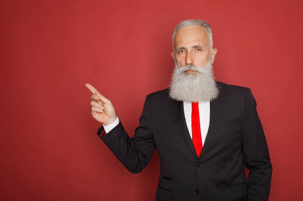 Copy space at his hand. Bearded senior man in suit and tie pointing copy space and smiling while standing against red background - Photo, image