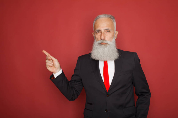 Copy space at his hand. Bearded senior man in suit and tie pointing copy space and smiling while standing against red background - Фото, зображення