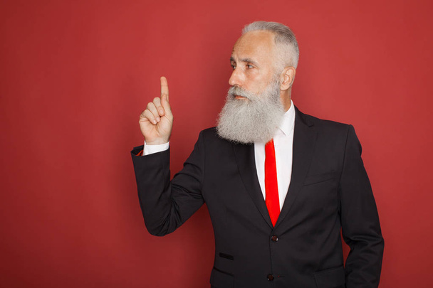 Copy space at his hand. Bearded senior man in suit and tie pointing copy space and smiling while standing against red background - Photo, image
