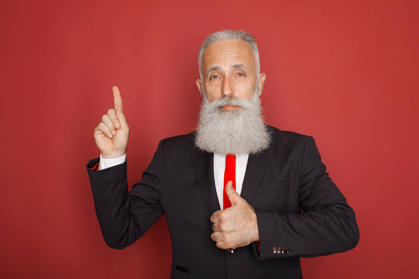 Copy space at his hand. Bearded senior man in suit and tie pointing copy space and smiling while standing against red background - 写真・画像
