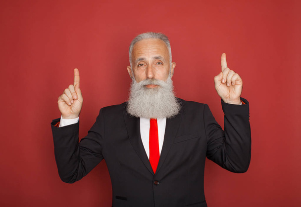Copy space at his hand. Bearded senior man in suit and tie pointing copy space and smiling while standing against red background - Foto, imagen