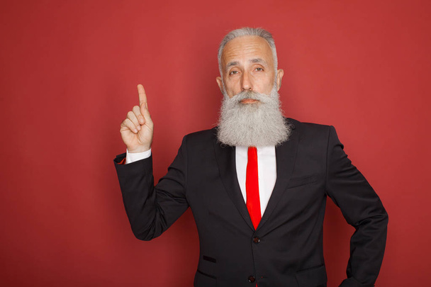 Copy space at his hand. Bearded senior man in suit and tie pointing copy space and smiling while standing against red background - Фото, изображение