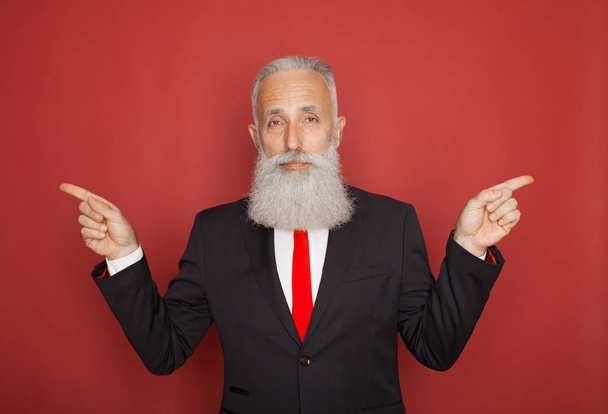 Copy space at his hand. Bearded senior man in suit and tie pointing copy space and smiling while standing against red background - Foto, Imagen