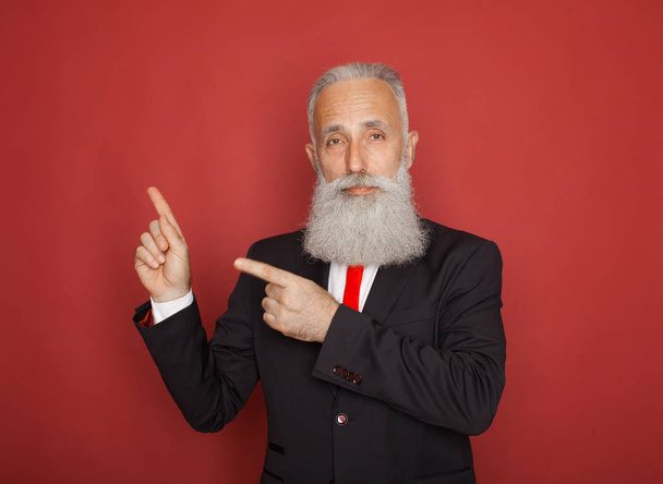 Copy space at his hand. Bearded senior man in suit and tie pointing copy space and smiling while standing against red background - Photo, Image