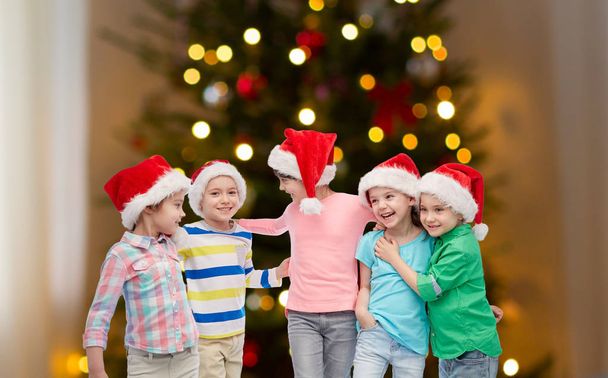 childhood, holidays and people concept - group of happy smiling little children in santa hats hugging over christmas tree lights on background - Foto, Imagem