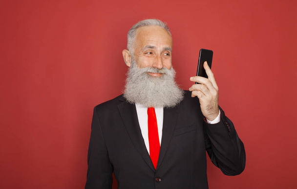 Studio shot of happy senior bearded businessman smiling while using mobile phone against red background - Foto, Imagem