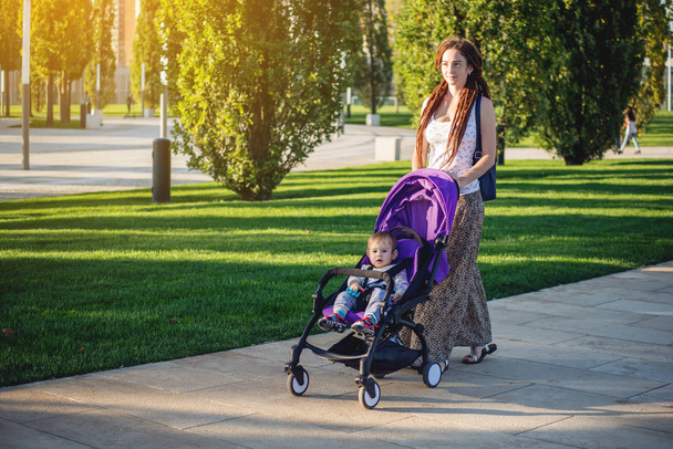 Young modern mom with baby son in stroller walking in Sunny Park. The concept of the joy of motherhood and autumn mood - 写真・画像