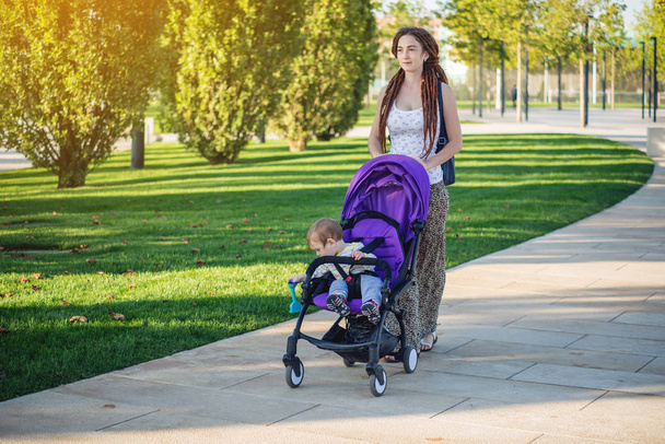 Young modern mom with baby son in stroller walking in Sunny Park. The concept of the joy of motherhood and autumn mood - Photo, Image