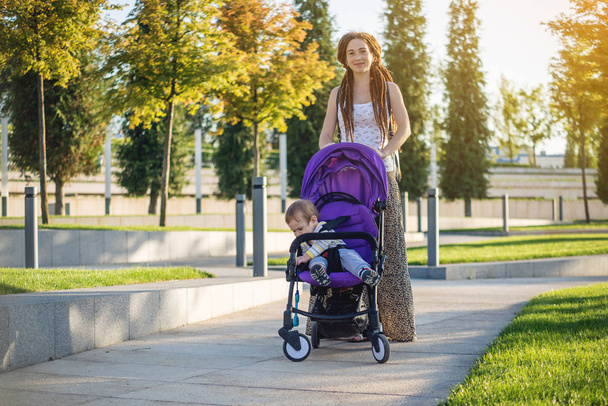 Young modern mother with baby son in stroller walking in Sunny Park. The concept of the joy of motherhood and autumn mood - Photo, Image