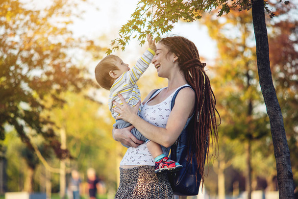 Young modern happy mom with baby son walking in Sunny Park. The concept of the joy of motherhood and autumn mood - Фото, изображение