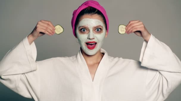 A girl with a mask on her face and a towel on her head covers her eyes with cucumbers on a gray background. A woman takes care of her face with a cosmetic mask. The concept of cosmetology. - Πλάνα, βίντεο