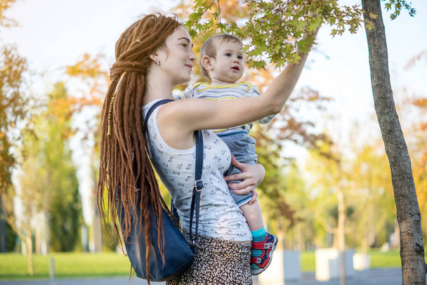 Jovem mãe feliz moderna com bebê filho andando em Sunny Park. O conceito da alegria da maternidade e do clima de outono
 - Foto, Imagem
