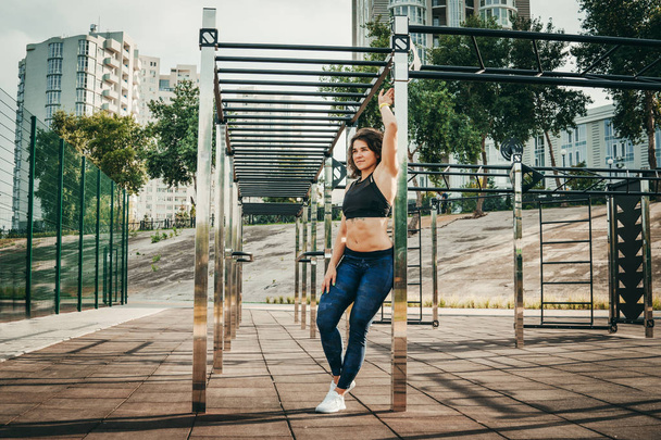 The theme women sports and health. Beautiful sexy caucasian woman with curly long hair posing on outdoor sports ground holding horizontal bar with tattoo on stomach in sports top and tight pants - Photo, Image
