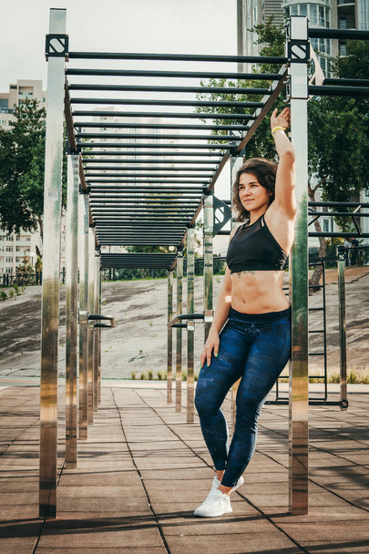 The theme women sports and health. Beautiful sexy caucasian woman with curly long hair posing on outdoor sports ground holding horizontal bar with tattoo on stomach in sports top and tight pants - 写真・画像