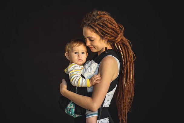 Portré fiatal boldog anya és a baba fiú ergo hátizsák, a háttér sötét falon. A koncepció a modern szülők - Fotó, kép