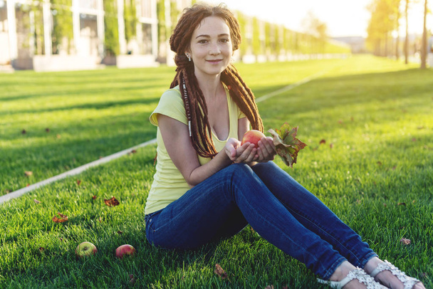 Modern, fiatal lány a parkban, egy zöld rét, étkezési alma, és élvezi a napsütéses őszi hangulat a frizura raszta - Fotó, kép