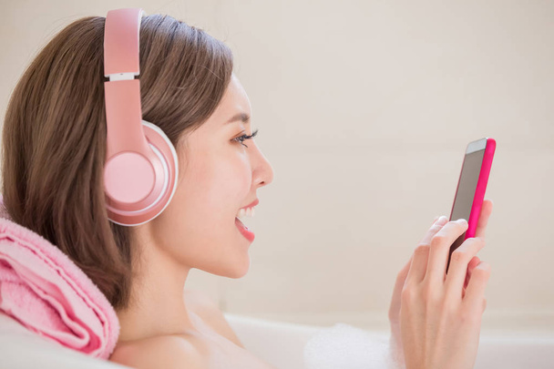 woman using  phone and wearing  headphones  in bathtub - Photo, Image