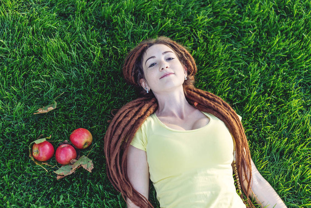 Modern, fiatal lány a parkban, egy zöld rét, étkezési alma, és élvezi a napsütéses őszi hangulat a frizura raszta - Fotó, kép