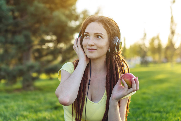 Beautiful modern woman listening to music with her headphones on the background of nature in autumn Sunny Park. The concept of Melomania and good mood - Photo, Image