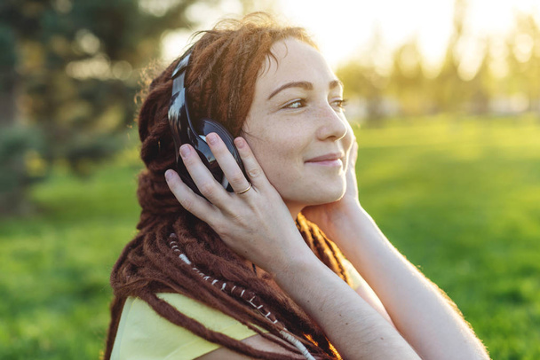 美しい現代の女性は、日当たりの良い公園の秋の自然の背景に彼女のヘッドフォンで音楽を聴きます。Melomania と良い気分のコンセプト - 写真・画像