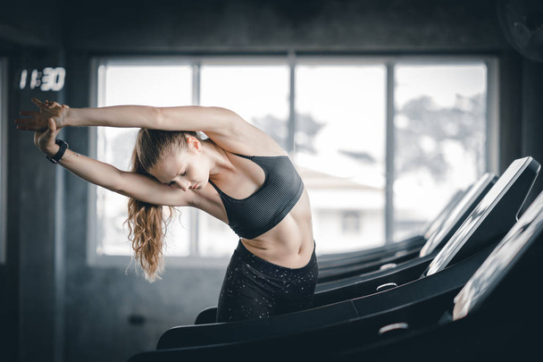 Jovem mulher treino de fitness atraente no ginásio. Mulher alongando os músculos e relaxando após o exercício na esteira da máquina no clube de fitness
. - Foto, Imagem