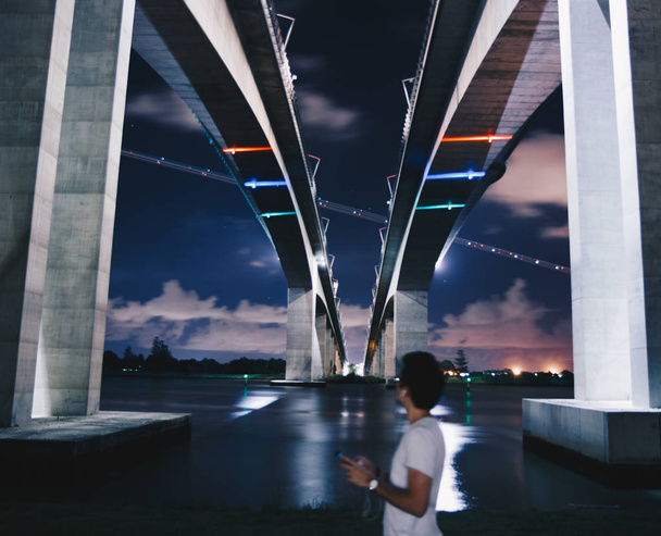 Onderstaande uitzicht op de brug van de snelweg en zijaanzicht van persoon met smartphone, oortelefoons permanent op rivier wal in Brisbane, Australië - Foto, afbeelding