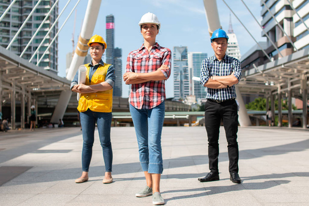 Trzy inżynierii bezpieczeństwa inżynier nosić kask stojąc z rękami skrzyżowanymi przytrzymując kontroli tablet i uśmiech na budynku poza godzinami. Narzędzia projektowe i budowlane koncepcja. - Zdjęcie, obraz