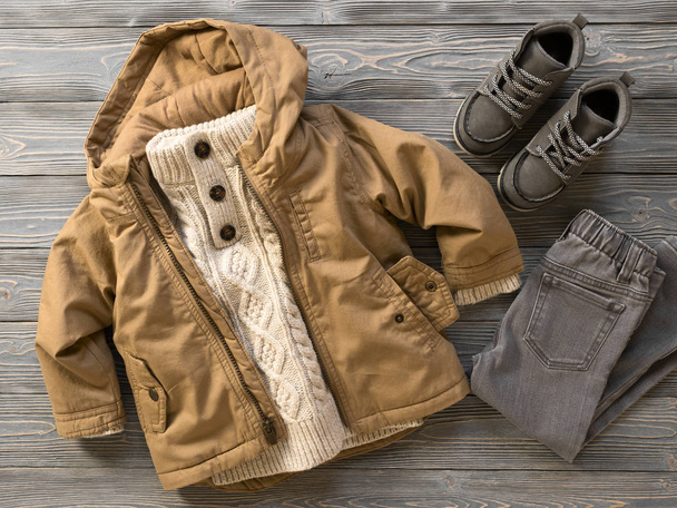Μόδα παιδικά ρούχα, παπούτσια (δεμένη πουλόβερ, μπουφάν με κουκούλα καμβά, τζιν, σουέτ μπότες). Ντύσιμο για αγοράκι. Χειμώνας, φθινόπωρο συλλογή. Οργανικό βαμβάκι. Το Top view, επίπεδη lay - Φωτογραφία, εικόνα