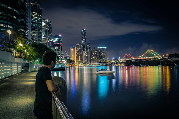 Boční pohled na člověka při pohledu na jachtě na vodní plocha poblíž osvětlenými mrakodrapy na břehu v noci v Brisbane, Austrálie - Fotografie, Obrázek