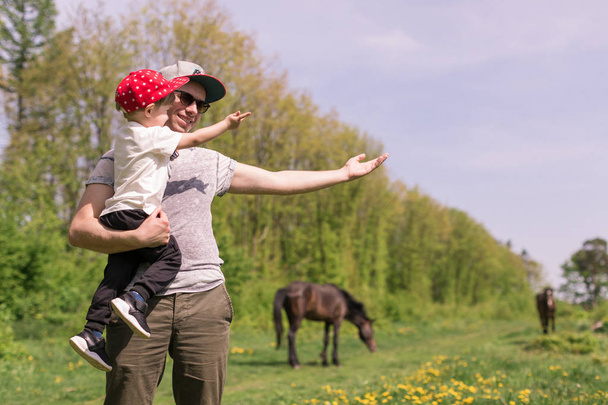 The young happy father standing on a clearing where the horses are grazing and holding the cute little son in arms and showing something him - Fotoğraf, Görsel