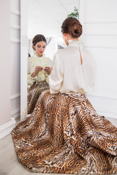 The beautiful, stylish young woman sitting in front of the mirror and holding an earring in hand indoors - Photo, image