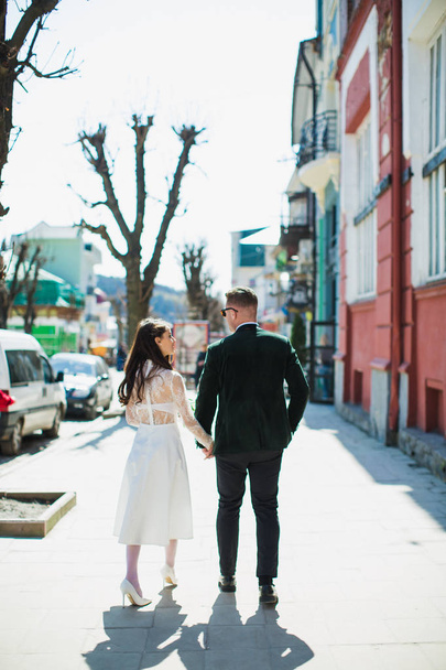 A side view of romantic young stylish newlyweds who are walking and holding their hands outdoors - Fotó, kép