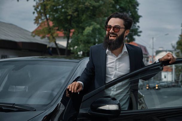 Beautiful, stylish, bearded man, smiling happy while standing near car, wearing a black jacket, white shirt and fashionable sunglasses. Portrait attractive man. Life lover. City people. - Photo, Image