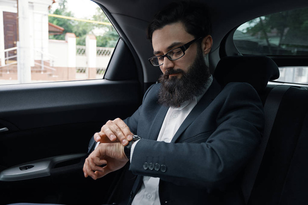 Je trouverai les meilleures solutions. Homme d'affaires confiant réfléchissant à l'efficacité et à la croissance de l'entreprise tout en regardant sa montre, assis sur le siège arrière d'une voiture
. - Photo, image