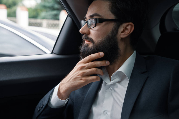I will find the best solutions. Confident businessman thinking about efficiency and company growth while touching his beard and looking through the window, sitting on the back seat of a car. - Photo, Image