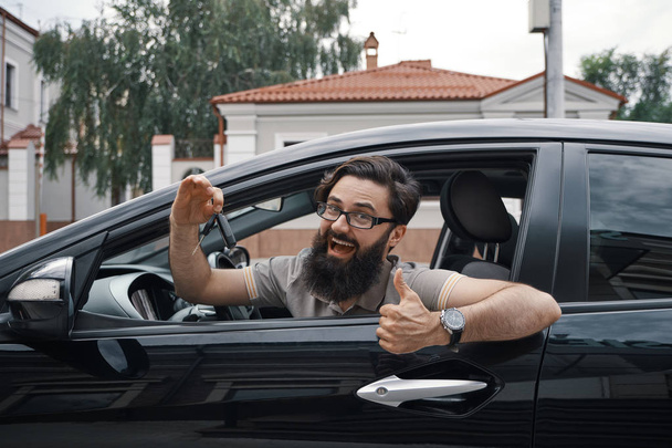 Автомобільний дилер, найщасливіший клієнт. Чоловік тримає ключі від машини, показуючи великі пальці вгору і посміхаючись, з зубами, сидячи в машині, дивлячись на камеру через вікно
.. - Фото, зображення