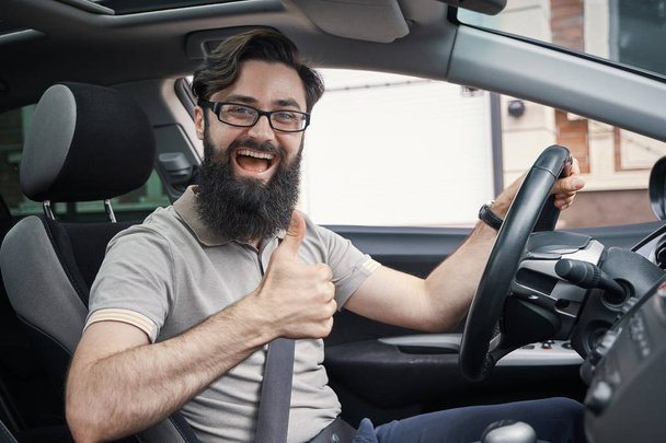 Οδηγός άνθρωπος ευτυχής χαμογελαστοί προβολή μπράβο αυτοκίνητο άθλημα με κτίρια της πόλης, στο παρασκήνιο. Όμορφος νεαρός άνδρας ενθουσιασμένος για το νέο όχημά του. Θετικό πρόσωπο έκφραση - Φωτογραφία, εικόνα