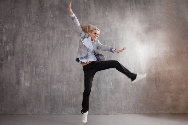 Young blonde woman in business suit and sneakers jumping for joy, gray textured background - Valokuva, kuva