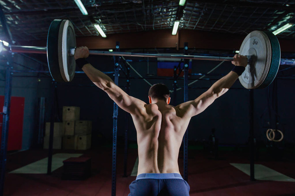 Portrait of a handsome athlete from behind. Athlete raises the barbell over your head. Studio shots in the dark tone. Cross style fit, deadlift - Photo, image