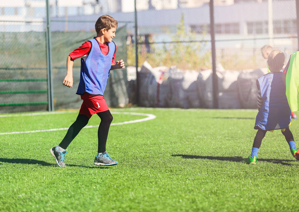 cute young boy in red and blue uniform plays football with his team on the green grass. Team game, training, active lifestyle, hobby, sport for kids concept - 写真・画像