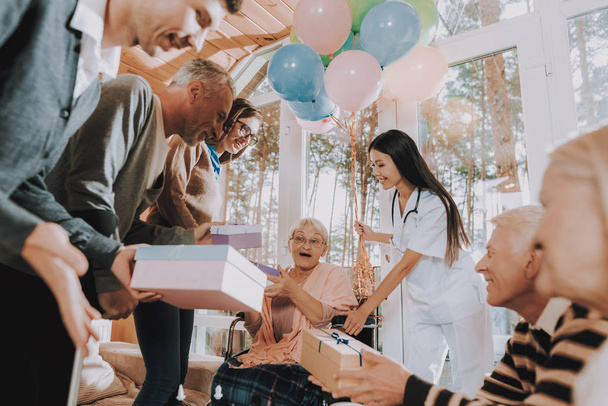 Gifts. Wheelchair. Madly Happy Retiree. Birthday Party. Celebration. Balloons. Guests. Young and Old People. Nursing Home. Congratulate. Elderly Woman. Give Gifts. Very Surprised. Very Happy. - Photo, Image