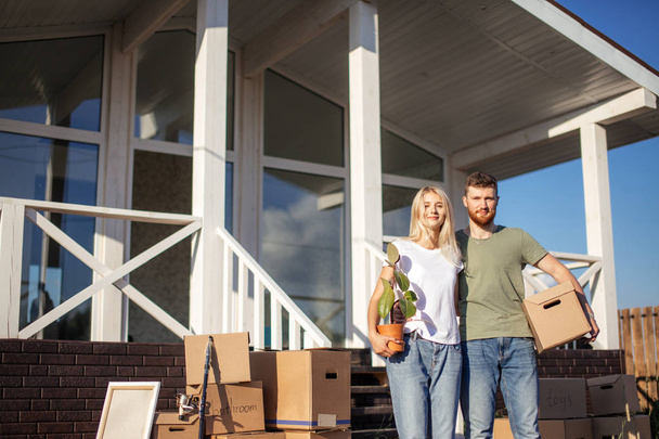 Husband and wife standing in front of new buying home with boxes - Фото, изображение