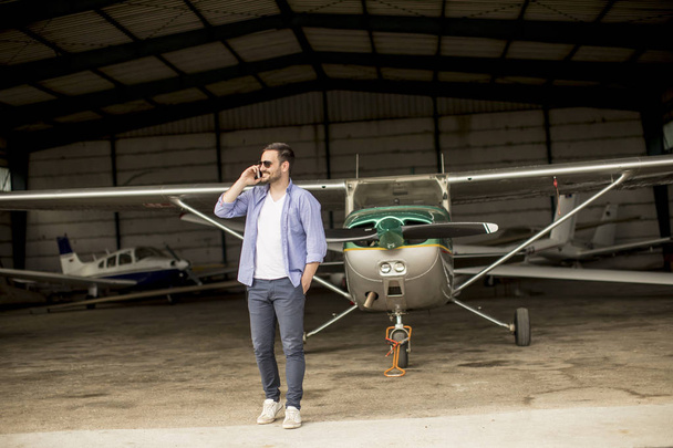 Jovem e bonito jovem piloto verificando avião no hangar e usando telefone celular
 - Foto, Imagem
